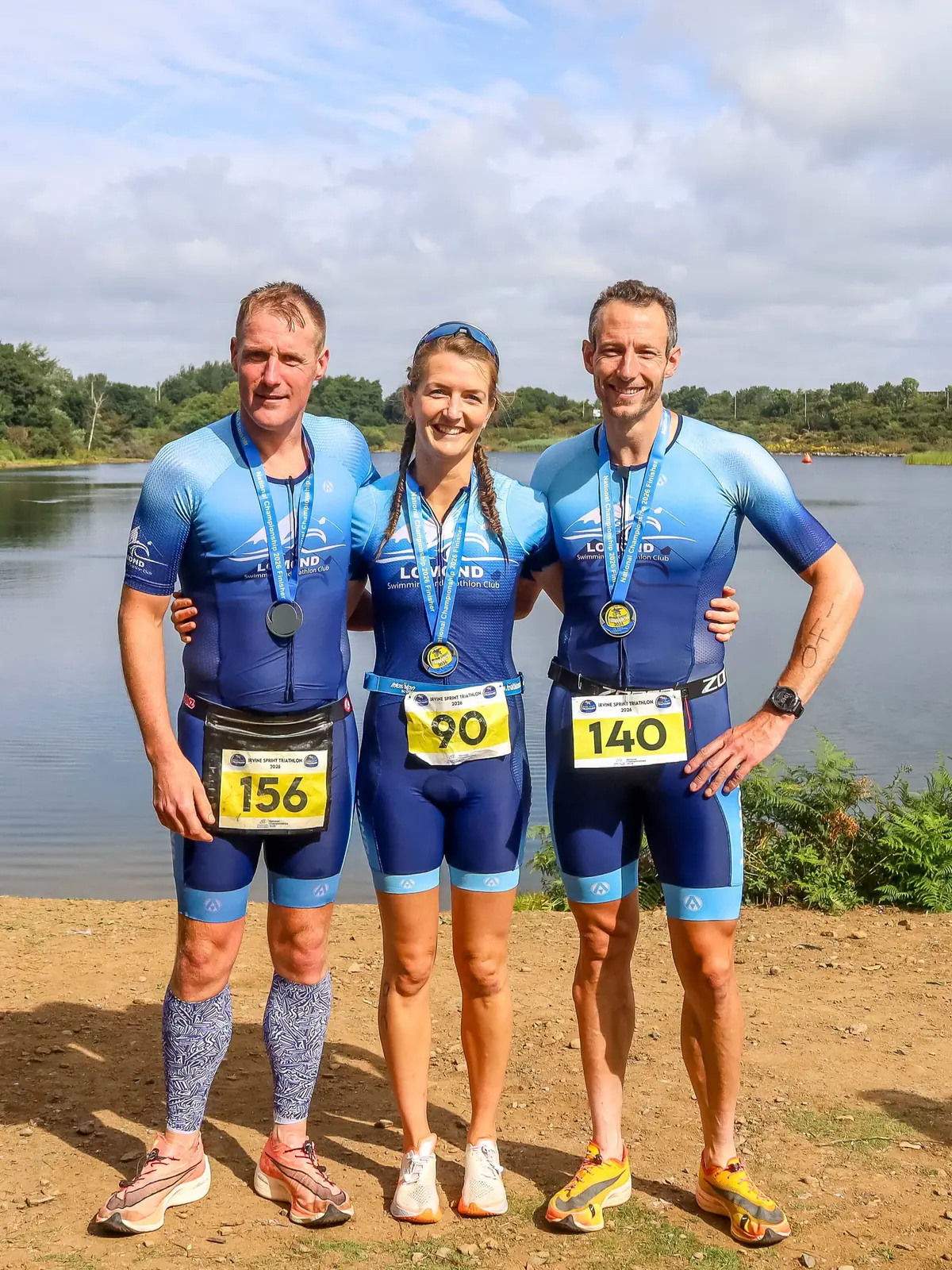 Three Lomond members in club kit with their finishers' medals beside the water